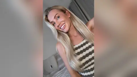 Smiling blonde woman in a striped knit tank top relaxing in a bright bedroom setting.