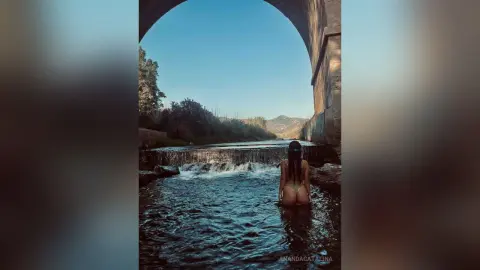 Young woman in a bikini relaxing in a river under a stone archway in a serene natural landscape.