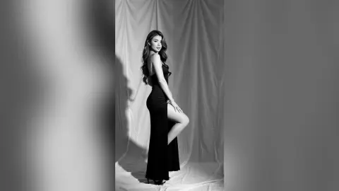 Elegant woman in a sleek black evening gown posing against draped fabric in a moody black and white studio portrait.