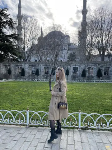 Blonde woman in a beige trench coat standing before the historic Blue Mosque in Istanbul on a cloudy day.
