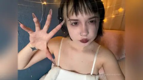 Young woman with short dark hair and nose ring wearing a white camisole in a cozy room with fairy lights.