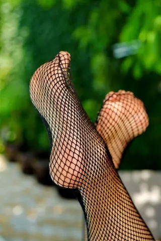 Close-up of feet wearing black fishnet stockings against a lush green garden background in bright sunlight.
