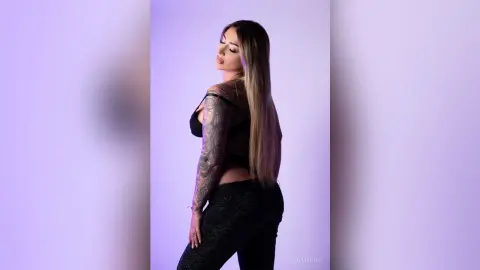 Elegant woman in a black mesh top and dark trousers posing confidently against a soft purple studio background.