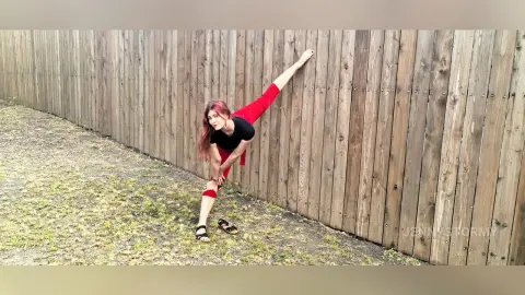 Redhead woman in black top and red shorts performing a graceful ballet stretch against a wooden fence.