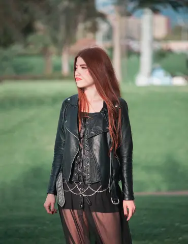 Redheaded woman in a black leather jacket and sheer skirt posing in a sunny green park.