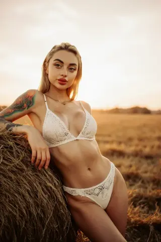 Blonde woman in white lace bikini leaning on hay bale in a golden sunset field.