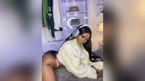 Glamorous woman in a white faux fur coat wearing headphones while relaxing in a modern bedroom.