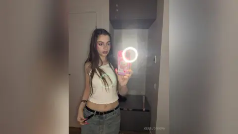 Young woman in a white tank top taking a selfie in a mirror with a ring light in a modern room.