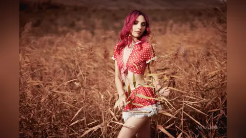 Woman with pink hair in a red polka dot crop top and skirt standing in a dreamy golden reed field.