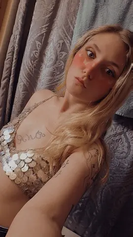 SummerBrookes poses in a gold sequin halter top, showing her long blonde hair and arm tattoos against a grey patterned curtain background.