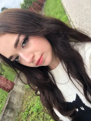 Close-up selfie of a young woman with long dark hair wearing a white sweater in a bright outdoor garden setting.