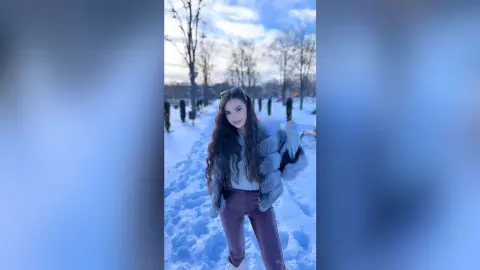 Young woman in a fuzzy grey coat and maroon trousers smiling in a snowy winter park under a blue sky.
