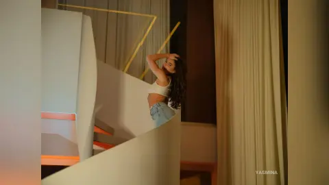 Stylish woman in a white crop top and denim shorts posing on a modern white staircase in a warm, elegant interior.