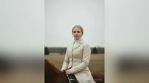 Elegant woman in a vintage cream equestrian outfit sitting on a brown horse in a misty field.
