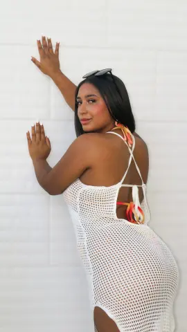Young woman in a white crochet mesh cover-up dress posing against a white brick wall in bright sunlight.