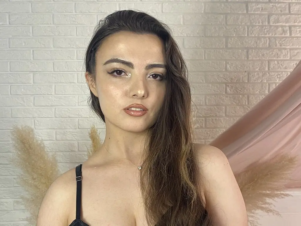 Young woman with long brown hair wearing a black strap top against a white brick wall and soft pink backdrop.