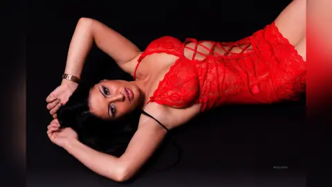 NicolleSun lies on a black background wearing a red lace bodysuit with a lace-up front, posing with her hands behind her head.