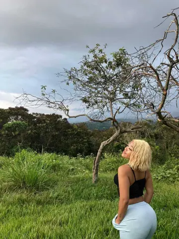 Blonde woman in a black crop top and light blue jeans standing in a lush green field under a cloudy sky.