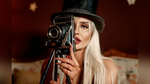 Gia peeks from behind a vintage black camera, her long blonde hair falling over her shoulders against a dark red background.