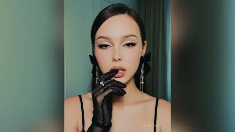TarenLeonetti poses with a hand to her lips, wearing black mesh gloves, large black bows, and dangling earrings against a teal background.