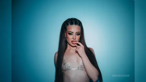 GracieCarter poses against a blue background wearing a silver glittery bra and letting her long dark hair hang down her shoulders.
