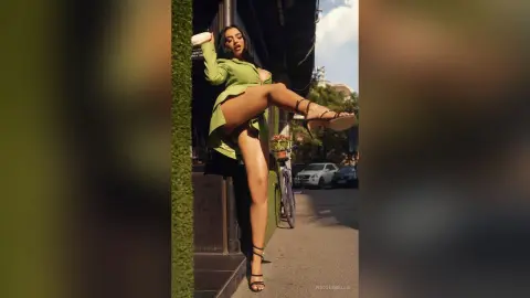 NicoleBella poses outdoors in a short green dress, lifting one leg to show her bare thigh and black strappy sandals.