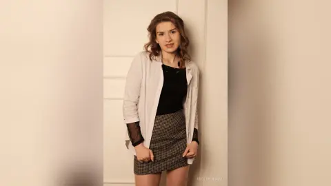 Smiling young woman in a white button-down shirt and plaid skirt posing against a white door.