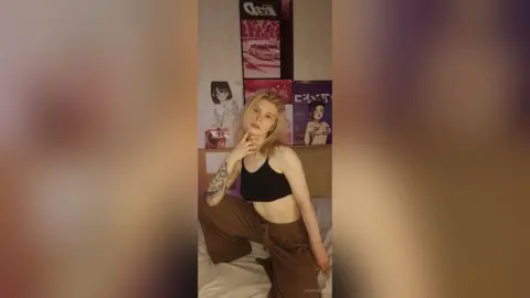 Young woman in a black crop top and brown trousers posing in a bedroom with anime posters.