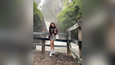 Woman in a brown leather jacket and grey skirt standing on a wooden bridge by a misty waterfall.
