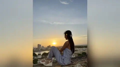 Young woman in striped jumpsuit sitting on a rooftop overlooking a city skyline at sunset.
