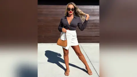 Stylish woman in a black blouse and white mini skirt posing confidently outdoors against a wooden wall.