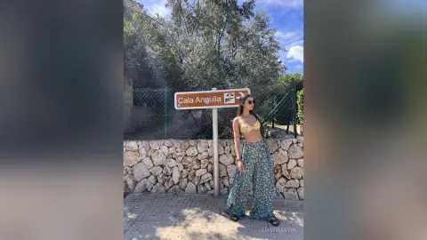 LucyMartin stands outdoors in front of a brown sign and stone wall wearing a yellow lace bra and blue patterned wide-leg trousers.