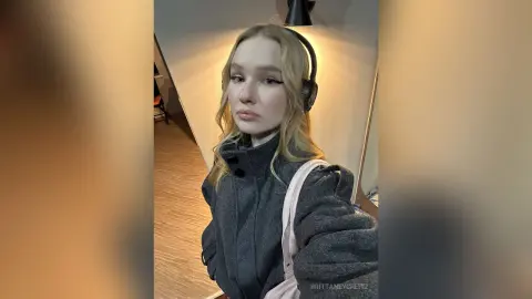 Blonde woman in a grey fleece jacket and headphones posing indoors with a casual, moody aesthetic.
