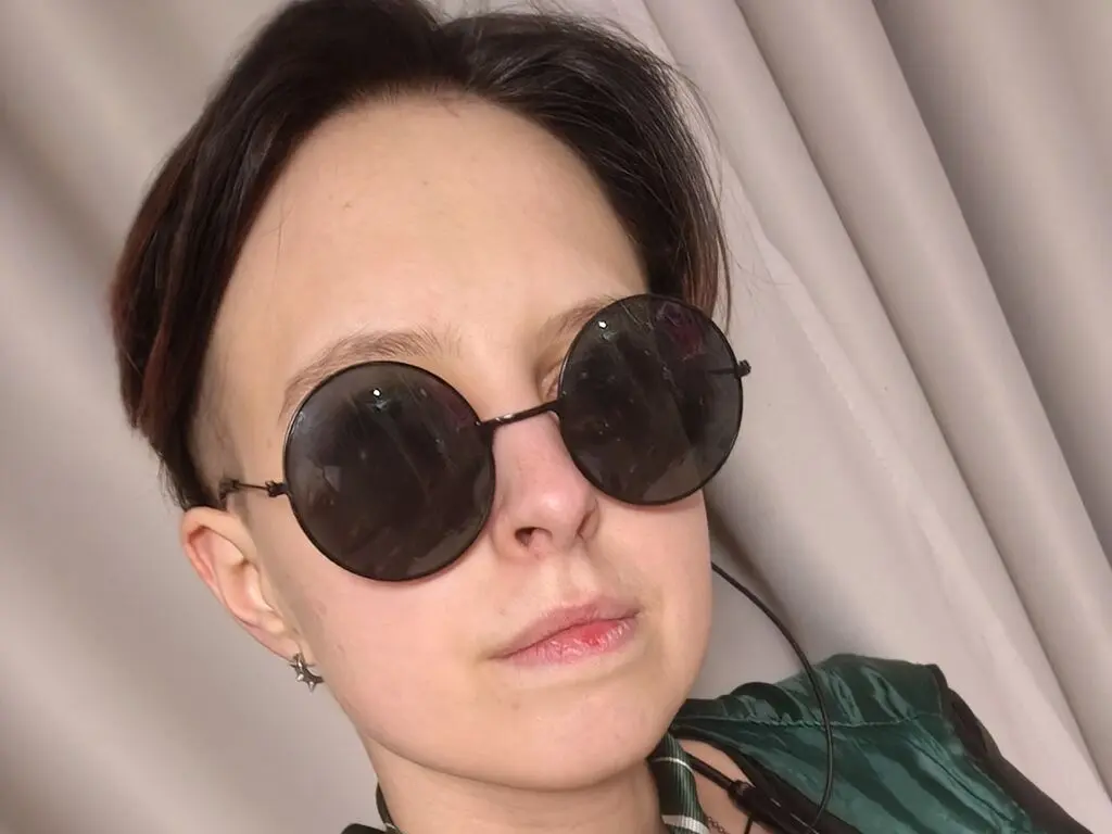 Young woman with short hair wearing round black sunglasses and green outfit in a bright indoor setting.