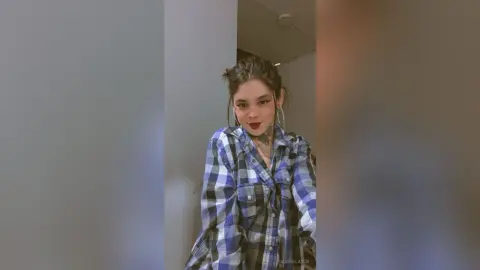Young woman with dark hair and tattoos wearing a blue plaid flannel shirt in a cozy indoor setting.