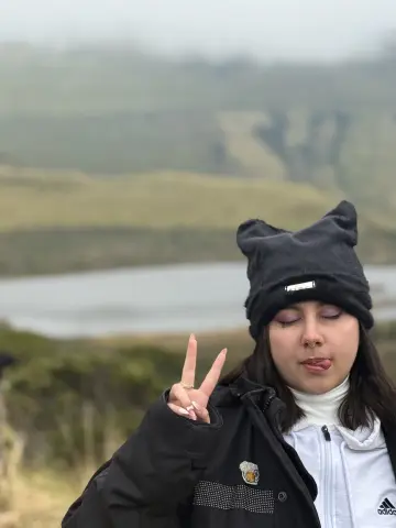 Young woman in a black beanie and white turtleneck making a peace sign in a misty mountain landscape.