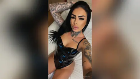 Tattooed woman in a black shiny bodysuit posing on a white bed with a confident mood.