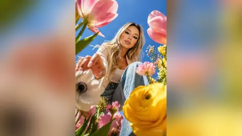Lucine poses outdoors in a field of pink and yellow flowers, wearing a white top and blue jeans with her long blonde hair down.