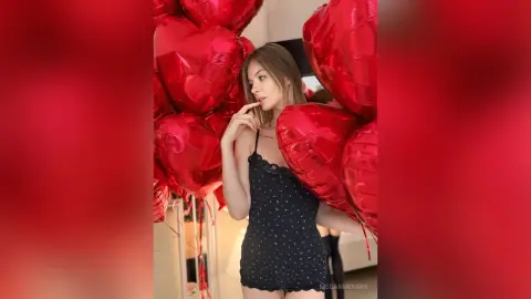 Young woman in a black floral slip dress posing with red heart balloons in a romantic indoor setting.