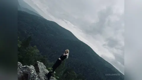 Blonde woman in black athletic wear standing on a rocky mountain peak under a moody, cloudy sky.