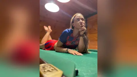 Young woman in a blue football jersey lying on a green pool table in a cozy wood-paneled room.