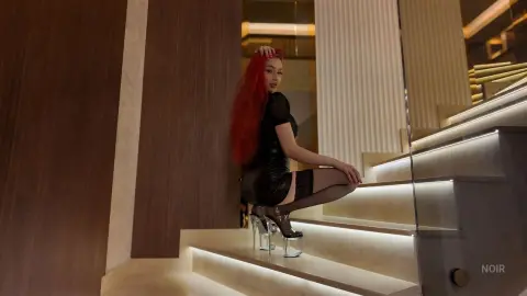 Red-haired woman in a black mini dress and platform heels sitting on modern lit stairs in a luxury interior.