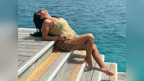 MissyJolie reclines on wooden deck steps overlooking the blue ocean, wearing a gold mesh bikini and gold waist chain.