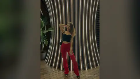 StacyHarland stands in front of a curved wooden slatted wall wearing a black tube top and red pants.