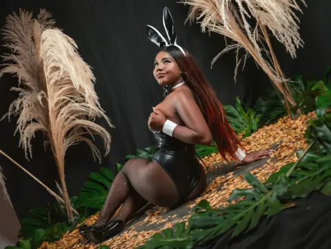 EvaJohhnss wears a black bunny outfit and ears while posing on a bed of leaves and pampas grass.