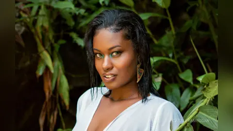 Beautiful Black woman in a white blouse smiling amidst lush green tropical foliage in a serene outdoor setting.