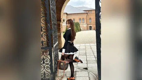 TiffanyAddams poses outdoors in a black and white schoolgirl-style outfit, holding a brown designer handbag while standing near a brick archway.