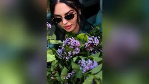 Stylish woman in sunglasses holding a large bouquet of purple lilacs in a moody, cinematic setting.