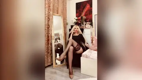 Glamorous blonde woman in a black dress and lace lingerie sitting in a luxurious gold and white bedroom.