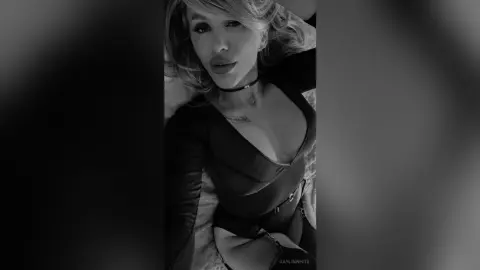Glamorous woman in a black bodysuit and choker relaxing in a moody, black and white indoor setting.